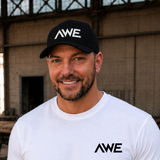 Man wearing a black cap and white t-shirt with 'AWE' logo in an industrial setting
